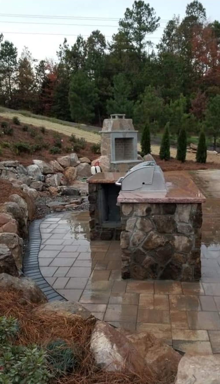 Outdoor patio with stone fireplace and pizza oven surrounded by landscaping, trees, and rock features on a sloped yard