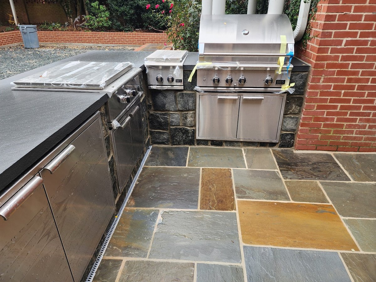 Outdoor kitchen with stainless steel grills and burners on a patio with mixed-color stone tiles against a brick wall