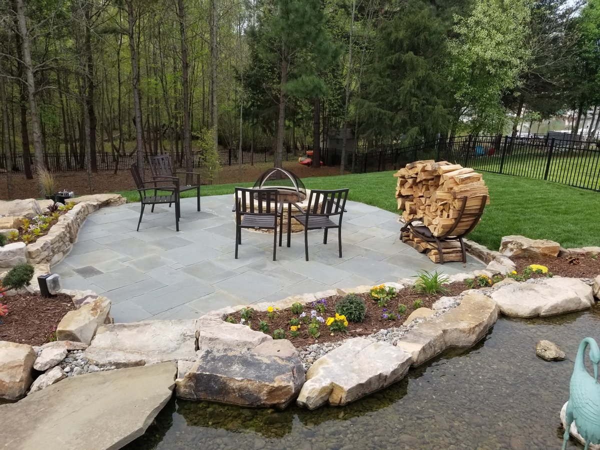 A stone patio with outdoor furniture and seating area surrounded by landscaping rocks and trees in a backyard setting