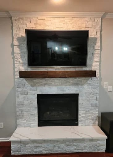 Modern fireplace with stacked stone surround, wall-mounted TV above wooden mantel, and marble hearth base